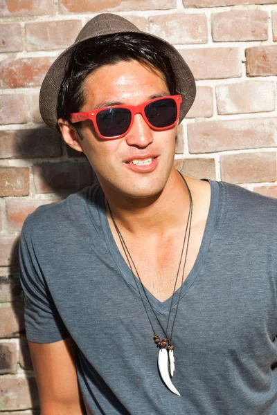 Αστικές ασιατικό άτομο με κόκκινα γυαλιά ηλίου. Ευπαρουσίαστο. ωραίος τύπος. φορώντας γκρι πουκάμισο και καπέλο. στέκεται μπροστά από τον τοίχο από τούβλα. — Φωτογραφία Αρχείου
