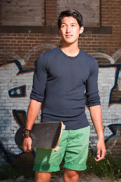 Hombre asiático urbano sosteniendo un monopatín. Muy guapo. Un tipo genial. Con camisa azul oscuro y pantalones cortos verdes. De pie frente a la pared de ladrillo con graffiti . —  Fotos de Stock