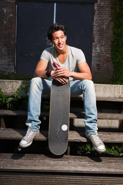 Ein urbaner asiatischer Mann mit Skateboard sitzt auf einer Treppe. gut aussehend. Cooler Typ. trägt graues Hemd und Jeans. altes, vernachlässigtes Gebäude im Hintergrund. — Stockfoto