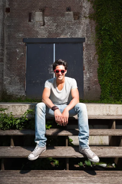 Urban uomo asiatico con occhiali da sole rossi seduti sulle scale. Bell'aspetto. Che figo. Indossa maglietta grigia e jeans. Vecchio edificio trascurato sullo sfondo . — Foto Stock