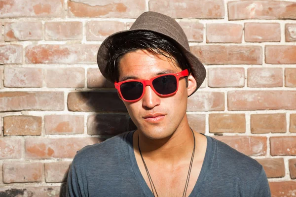 Urban asian man with red sunglasses. Good looking. Cool guy. Wearing grey shirt and hat. Standing in front of brick wall. — Stock Photo, Image