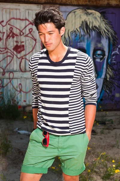 Urbano asiático homem. Boa aparência. Um tipo fixe. Vestindo camisola listrada branca azul e shorts verdes. Em pé na frente da parede de madeira com graffiti . — Fotografia de Stock
