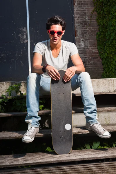 Urban uomo asiatico con occhiali da sole rossi e skateboard seduto sulle scale. Bell'aspetto. Che figo. Indossa maglietta grigia e jeans. Vecchio edificio trascurato sullo sfondo . — Foto Stock