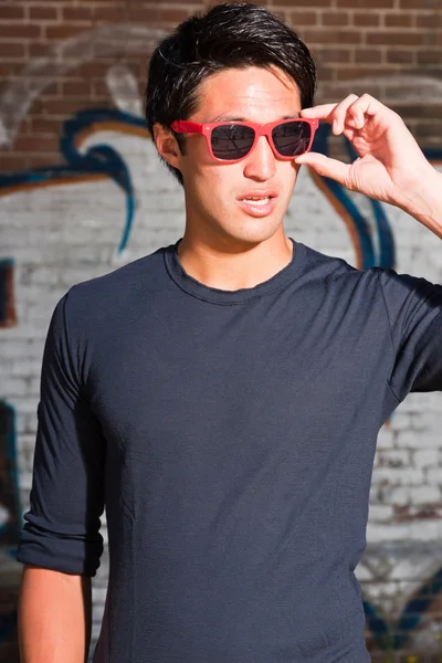 Hombre asiático urbano con gafas de sol rojas. Muy guapo. Un tipo genial. Llevando camisa azul oscuro. De pie frente a la pared de ladrillo con graffiti . —  Fotos de Stock
