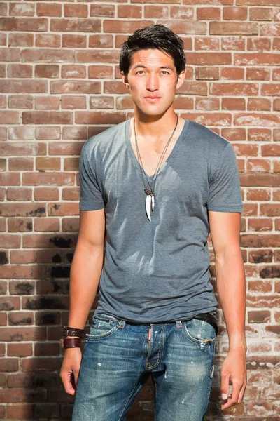Urbano asiático homem. Boa aparência. Um tipo fixe. Vestindo camisa cinza e jeans. Em pé na frente da parede de tijolo . — Fotografia de Stock