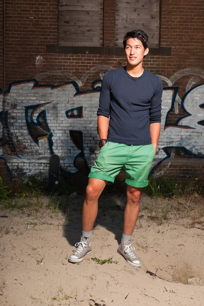 Urbano asiático homem. Boa aparência. Um tipo fixe. Vestindo camisa azul escuro e shorts verdes. Em pé na frente da parede de tijolo com graffiti . — Fotografia de Stock