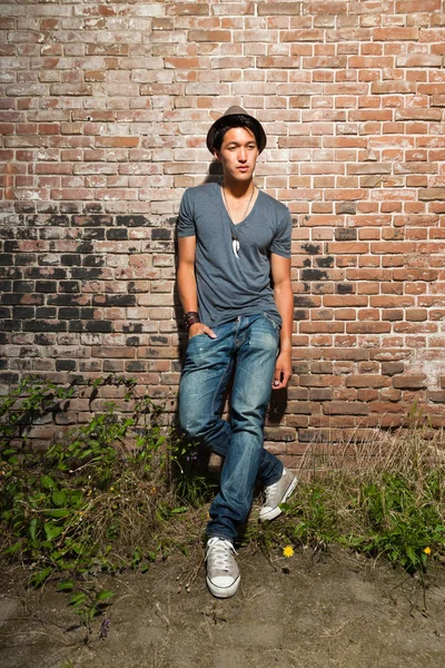 Urbano asiático homem. Boa aparência. Um tipo fixe. Vestindo camisa cinza e chapéu e jeans. Em pé na frente da parede de tijolo . — Fotografia de Stock