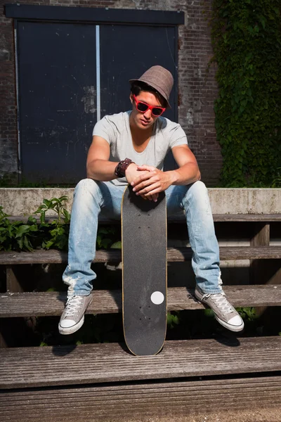Pria asia kota dengan topi, kacamata merah dan skateboard duduk di tangga. Tampan. Pria keren. Mengenakan kemeja abu-abu dan celana jeans. Bangunan lama yang diabaikan di latar belakang . — Stok Foto
