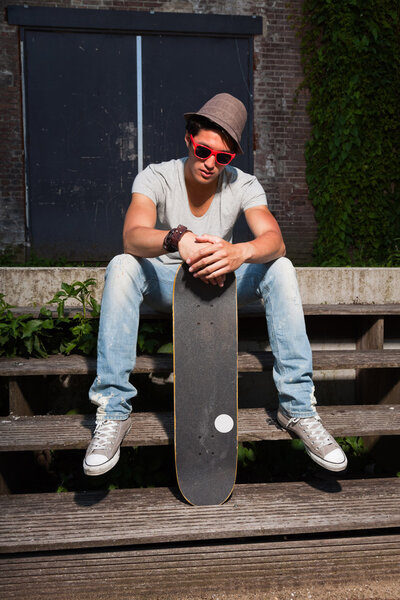 Urban asian man with hat, red sunglasses and skateboard sitting on stairs. Good looking. Cool guy. Wearing grey shirt and jeans. Old neglected building in the background.