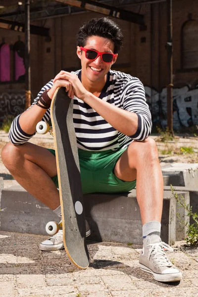 Hombre asiático urbano con gafas de sol rojas y monopatín sentado en la calle. Muy guapo. Un tipo genial. Usando jersey azul a rayas blancas y pantalones cortos verdes. Antiguo edificio abandonado en el fondo . — Foto de Stock