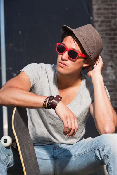 Stedelijke Aziatische man met hoed, rode zonnebril en skateboard zittend op trappen. Knap. koele kerel. het dragen van grijs shirt en spijkerbroek. oude verwaarloosd gebouw op de achtergrond. — Stockfoto