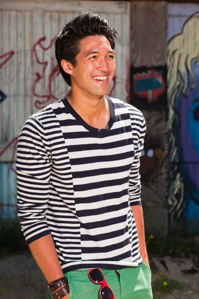 Urban asian man. Good looking. Cool guy. Wearing blue white striped sweater and green shorts. Standing in front of wooden wall with graffiti. — Stock Photo, Image