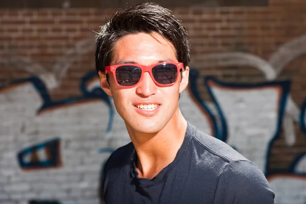 Urbano asiático homem com óculos de sol vermelhos. Boa aparência. Um tipo fixe. Vestindo camisa azul escuro. Em pé na frente da parede de tijolo com graffiti . — Fotografia de Stock