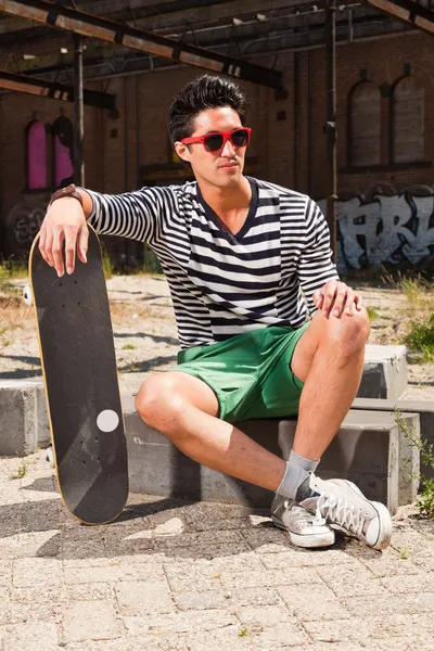 Городской азиат в красных солнцезащитных очках и скейтборде сидит на улице. Симпатичный. Крутой парень. В сине-белом полосатом свитере и зеленых шортах. Старое заброшенное здание на заднем плане . — стоковое фото