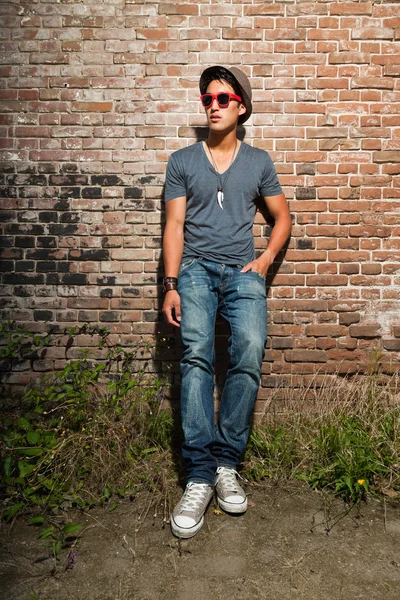 Hombre asiático urbano con gafas de sol rojas. Muy guapo. Un tipo genial. Usando camisa gris y sombrero y jeans. De pie frente a la pared de ladrillo . —  Fotos de Stock