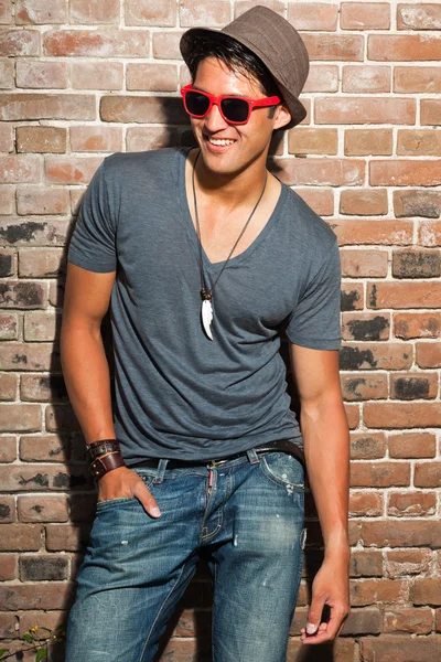 Hombre asiático urbano con gafas de sol rojas. Muy guapo. Un tipo genial. Usando camisa gris y sombrero y jeans. De pie frente a la pared de ladrillo . — Foto de Stock