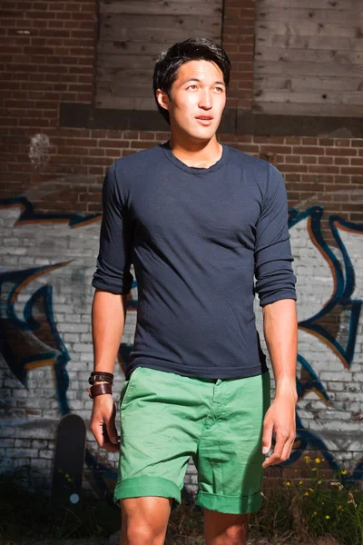 都市のアジア人男性。素敵ですね。クールな男。暗い青いシャツと緑のショート パンツを着ています。グラフィティでレンガの壁の前に立って. — ストック写真