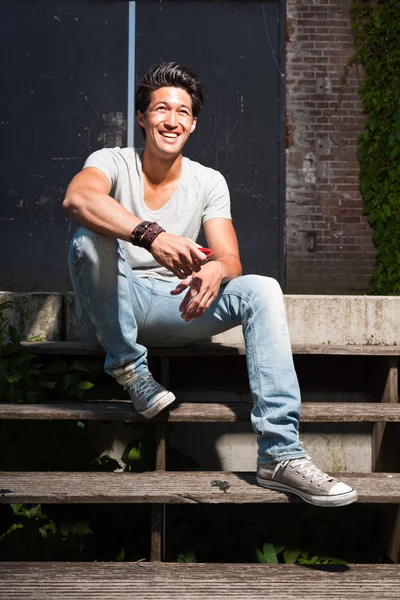 Urbano asiático homem sentado em escadas. Boa aparência. Um tipo fixe. Vestindo camisa cinza e jeans. Antigo edifício negligenciado em segundo plano . — Fotografia de Stock