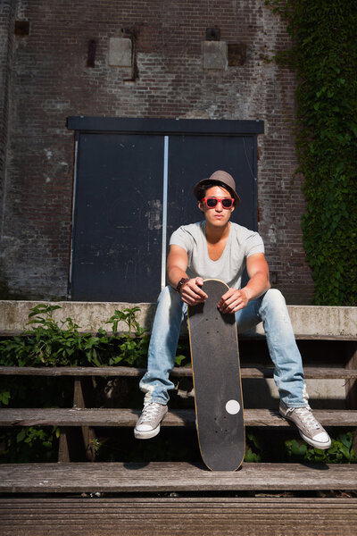 Urban asian man with hat, red sunglasses and skateboard sitting on stairs. Good looking. Cool guy. Wearing grey shirt and jeans. Old neglected building in the background.