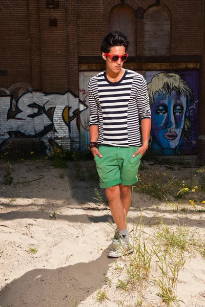 Urbano asiático homem com óculos de sol vermelhos. Boa aparência. Um tipo fixe. Vestindo camisola listrada branca azul e shorts verdes. Em pé na frente da parede com graffiti . — Fotografia de Stock