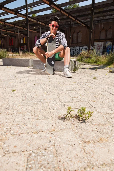 Urban uomo asiatico con occhiali da sole rossi e skateboard seduto sulla strada. Bell'aspetto. Che figo. Indossa maglione a righe bianche blu e pantaloncini verdi. Vecchio edificio trascurato sullo sfondo . — Foto Stock