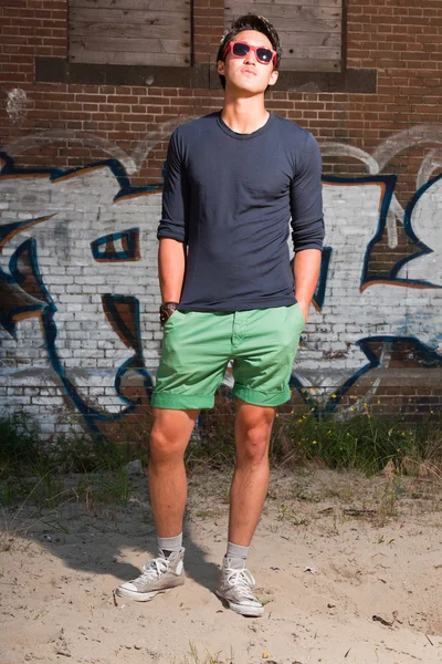 Urbano asiático homem com óculos de sol vermelhos. Boa aparência. Um tipo fixe. Vestindo camisa azul escuro e shorts verdes. Em pé na frente da parede de tijolo com graffiti . — Fotografia de Stock