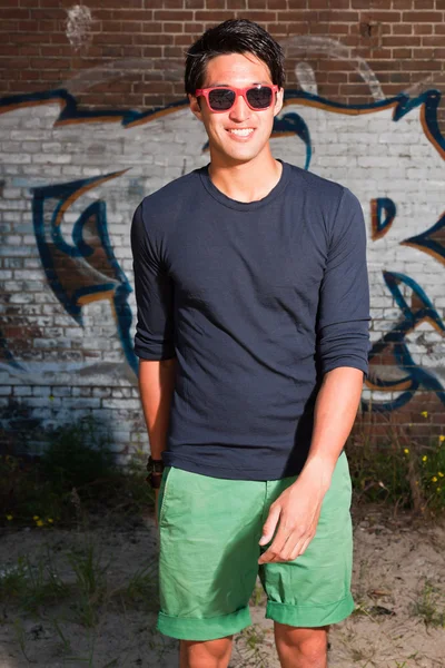 Αστικές ασιατικό άτομο με κόκκινα γυαλιά ηλίου. Ευπαρουσίαστο. ωραίος τύπος. φοράει σκούρο μπλε πουκάμισο και πράσινο σορτς. στέκεται μπροστά από τον τοίχο από τούβλα με γκράφιτι. — Φωτογραφία Αρχείου