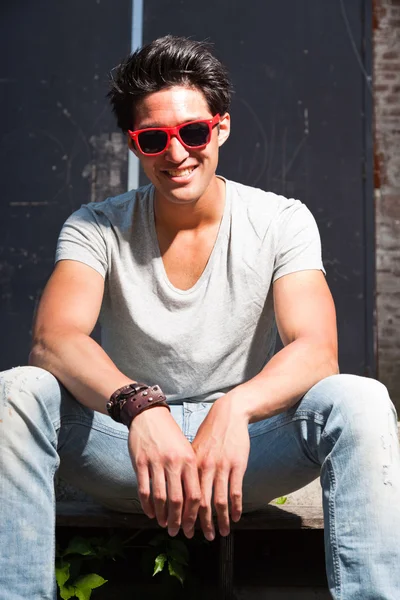 Urban uomo asiatico con occhiali da sole rossi seduti sulle scale. Bell'aspetto. Che figo. Indossa maglietta grigia e jeans. Vecchio edificio trascurato sullo sfondo . — Foto Stock