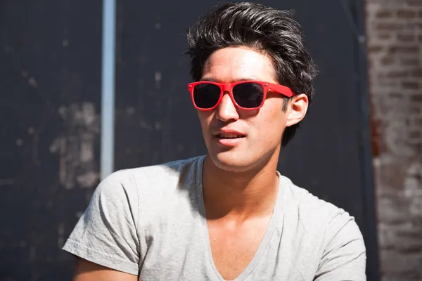 Urban asian man with red sunglasses. Good looking. Cool guy. Wearing grey shirt and jeans. Old neglected building in the background. — Stock Photo, Image