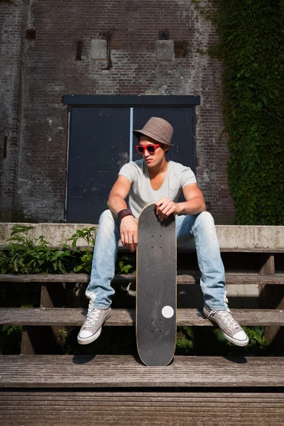 Hombre asiático urbano con sombrero, gafas de sol rojas y monopatín sentado en las escaleras. Muy guapo. Un tipo genial. Usando camisa gris y jeans. Antiguo edificio abandonado en el fondo . —  Fotos de Stock