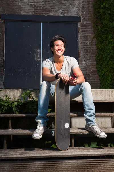 Městské Asiat s skateboard sedí na schodech. Krásné. cool chlapík. na sobě šedé tričko a džíny. staré zanedbané budově v pozadí. — Stock fotografie