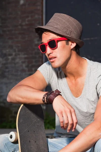 Hombre asiático urbano con sombrero, gafas de sol rojas y monopatín sentado en las escaleras. Muy guapo. Un tipo genial. Usando camisa gris y jeans. Antiguo edificio abandonado en el fondo . — Foto de Stock
