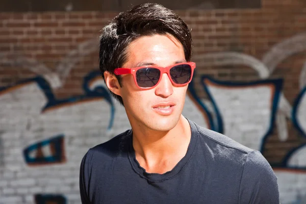 Hombre asiático urbano con gafas de sol rojas. Muy guapo. Un tipo genial. Llevando camisa azul oscuro. De pie frente a la pared de ladrillo con graffiti . — Foto de Stock