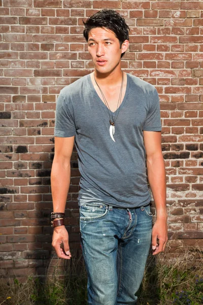 Městské Asiat. Krásné. cool chlapík. na sobě šedé tričko a džíny. stojící před cihlová zeď. — Stock fotografie