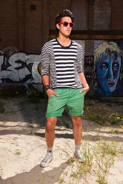 Αστικές ασιατικό άτομο με κόκκινα γυαλιά ηλίου. Ευπαρουσίαστο. ωραίος τύπος. φορώντας μπλε και λευκό ριγέ πουλόβερ και πράσινο σορτς. στέκεται μπροστά από τον τοίχο με γκράφιτι. — Φωτογραφία Αρχείου