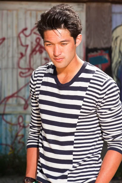 Hombre asiático urbano. Muy guapo. Un tipo genial. Usando jersey azul a rayas blancas. De pie frente a la pared de madera con graffiti . — Foto de Stock