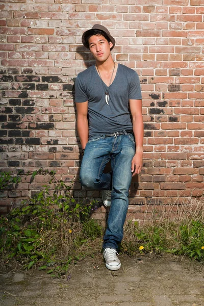 Homme asiatique urbain. Beau look. Cool mec. Il porte une chemise grise, un chapeau et un jean. Debout devant le mur de briques . — Photo