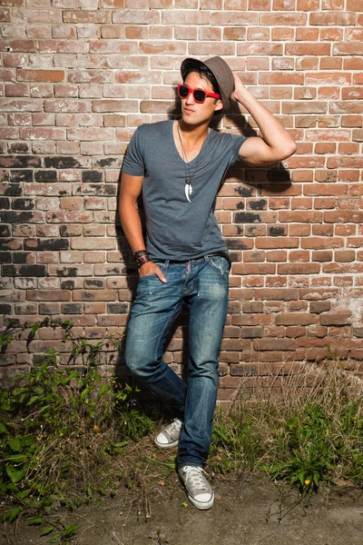 Urban asian man with red sunglasses. Good looking. Cool guy. Wearing grey shirt and hat and jeans. Standing in front of brick wall. — Stock Photo, Image