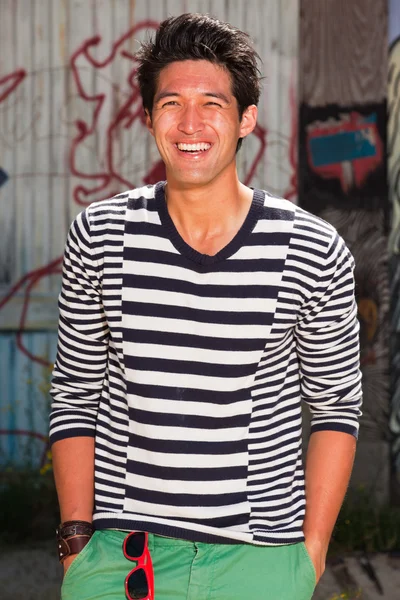 Hombre asiático urbano. Muy guapo. Un tipo genial. Usando jersey azul a rayas blancas y pantalones cortos verdes. De pie frente a la pared de madera con graffiti . —  Fotos de Stock