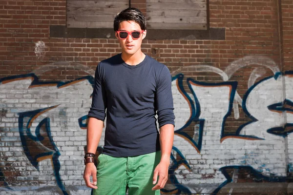 Hombre asiático urbano con gafas de sol rojas. Muy guapo. Un tipo genial. Con camisa azul oscuro y pantalones cortos verdes. De pie frente a la pared de ladrillo con graffiti . — Foto de Stock