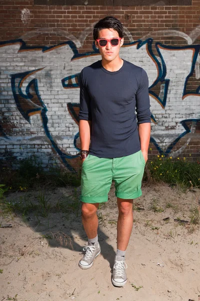 Αστικές ασιατικό άτομο με κόκκινα γυαλιά ηλίου. Ευπαρουσίαστο. ωραίος τύπος. φοράει σκούρο μπλε πουκάμισο και πράσινο σορτς. στέκεται μπροστά από τον τοίχο από τούβλα με γκράφιτι. — Φωτογραφία Αρχείου