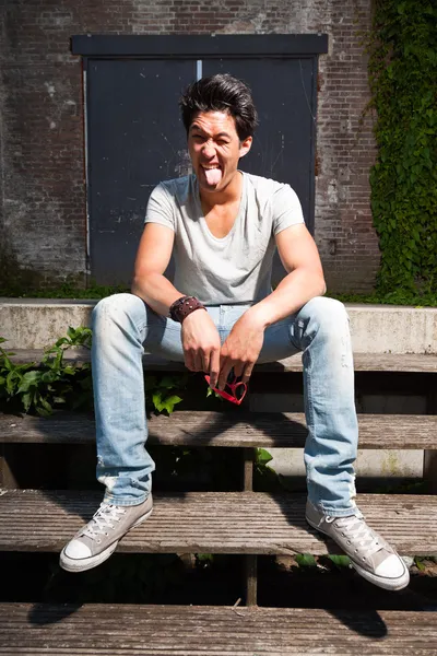 Urbano asiatico uomo making divertente faccia seduta su scale. Bell'aspetto. Che figo. Indossa maglietta grigia e jeans. Vecchio edificio trascurato sullo sfondo . — Foto Stock