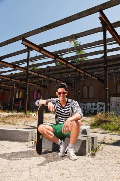 Urban uomo asiatico con occhiali da sole rossi e skateboard seduto sulla strada. Bell'aspetto. Che figo. Indossa maglione a righe bianche blu e pantaloncini verdi. Vecchio edificio trascurato sullo sfondo . — Foto Stock