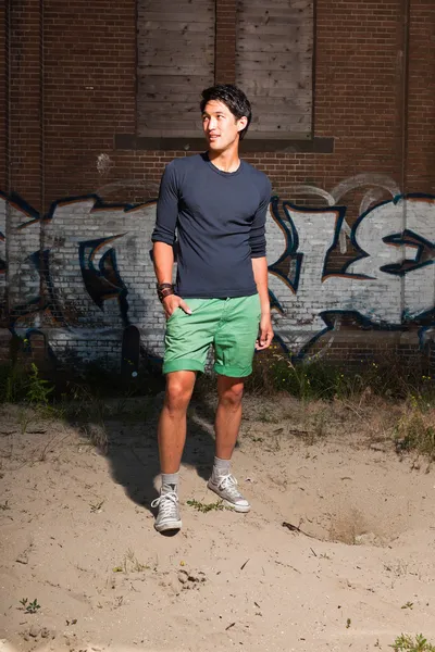 Urban asian man. Good looking. Cool guy. Wearing dark blue shirt and green shorts. Standing in front of brick wall with graffiti.