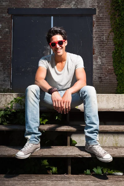 Hombre asiático urbano con gafas de sol rojas sentado en las escaleras. Muy guapo. Un tipo genial. Usando camisa gris y jeans. Antiguo edificio abandonado en el fondo . — Foto de Stock