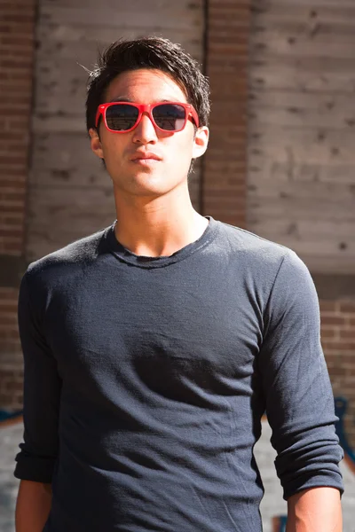 Homme asiatique urbain avec des lunettes de soleil rouges. Beau look. Cool mec. Elle porte une chemise bleu foncé. Debout devant un mur de briques avec des graffitis . — Photo