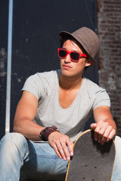 Urban asian man with hat, red sunglasses and skateboard sitting on stairs. Good looking. Cool guy. Wearing grey shirt and jeans. Old neglected building in the background.