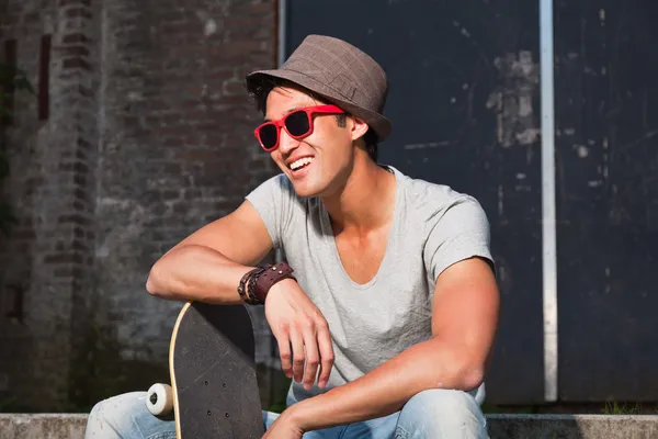 Hombre asiático urbano con sombrero, gafas de sol rojas y monopatín sentado en las escaleras. Muy guapo. Un tipo genial. Usando camisa gris y jeans. Antiguo edificio abandonado en el fondo . — Foto de Stock