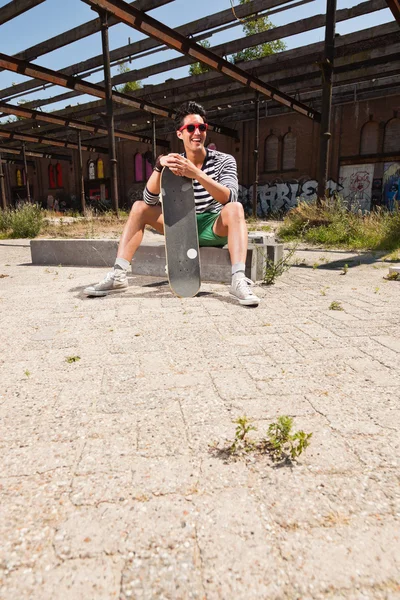 Αστικές ασιατικό άτομο με κόκκινα γυαλιά ηλίου και skateboard κάθεται στο δρόμο. Ευπαρουσίαστο. ωραίος τύπος. φορώντας μπλε και λευκό ριγέ πουλόβερ και πράσινο σορτς. παλιά παραμεληθεί κτίριο στο παρασκήνιο. — Φωτογραφία Αρχείου