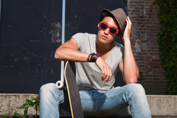 Urban asian man with hat, red sunglasses and skateboard sitting on stairs. Good looking. Cool guy. Wearing grey shirt and jeans. Old neglected building in the background.